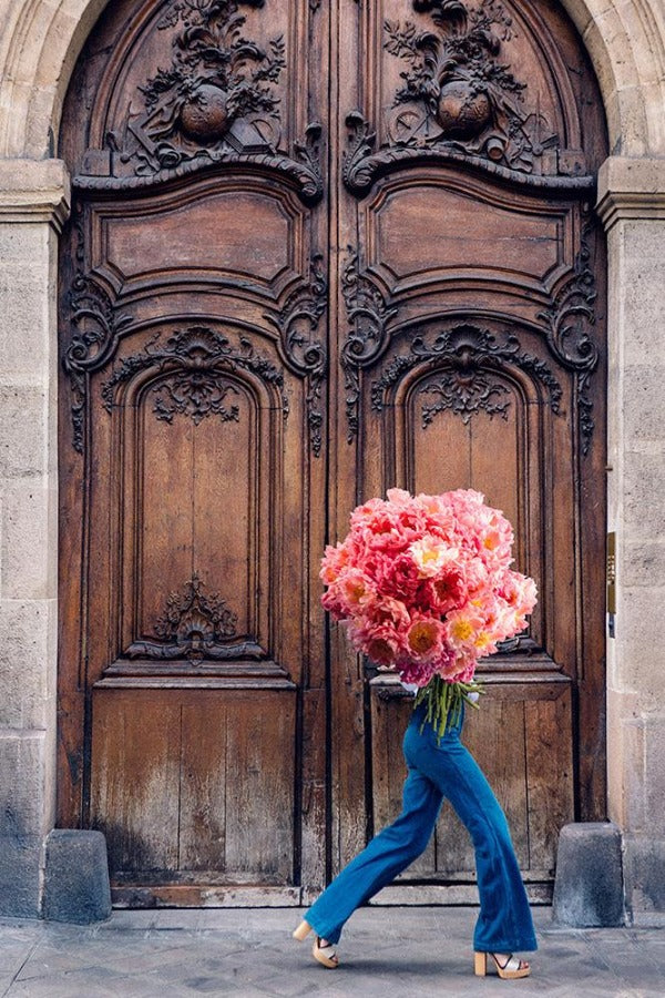 Breathtaking is a photo of a girl with a giant bouquet of coral peonies in front of a large wooden door in St Germain des Prés in Paris and is part of a limited edition series named Young Girl in Bloom by photographer Carla Coulson celebrating women loving and believing in themselves and building their self esteem by trusting their intuition.