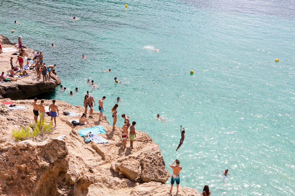 Freefall Ibiza, Carla Coulson Beach Photography fine art print, image of people on a rock ledge preparing to jump into the clear blue waters of Ibiza